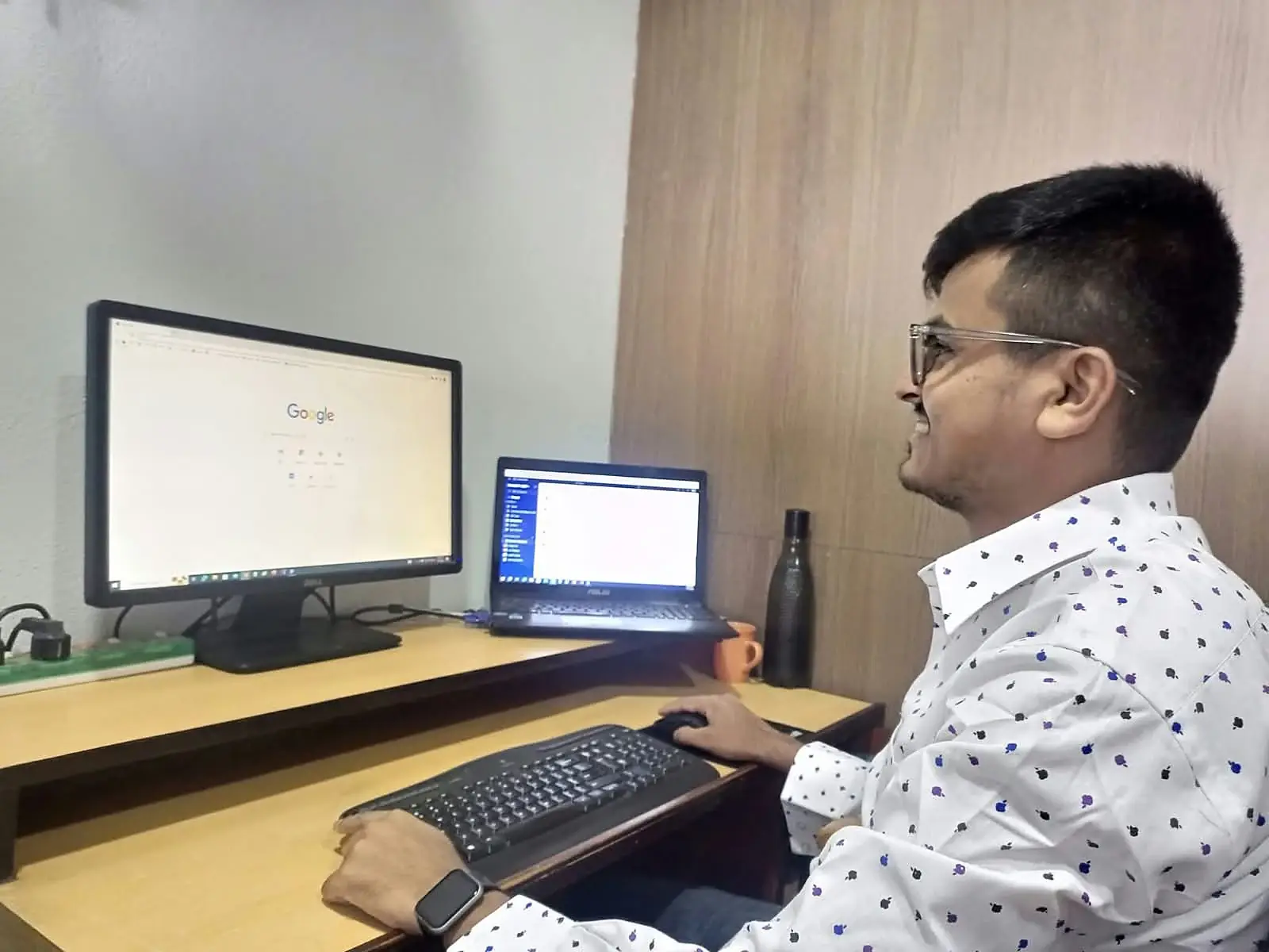 Hemraj smiling at his desk with a monitor and laptop
